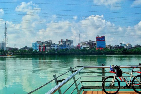 A white mountain bike with a blue helmet leaning on a lakeside railing, Dhaka skyline across the water.