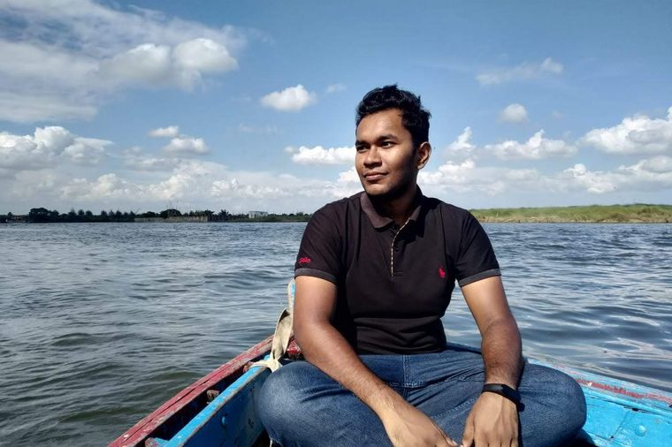 Yusuf sitting cross-legged in a wooden boat in the middle of a wide river, looking into the distance.