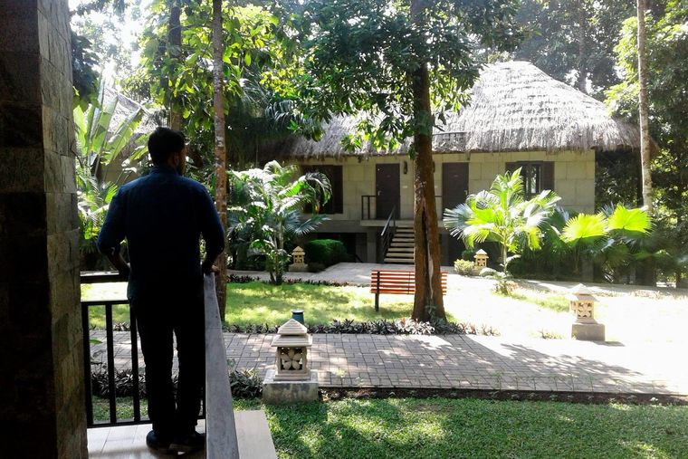 Yusuf standing on a balcony from behind, looking out over a leafy resort garden and a thatched-roof cottage.