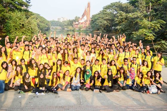 A big crowd of volunteers in yellow shirts cheering with raised arms beside a lake in Dhaka.