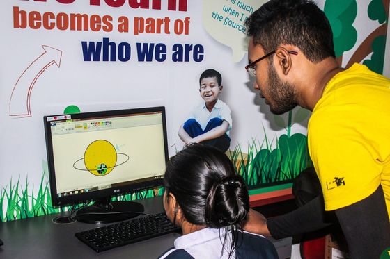 Yusuf in a yellow volunteer shirt guiding a young student who is drawing on a computer.